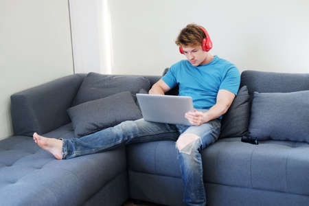 Young Caucasian man listening to musice by headphone and searching online social media with laptop in living room. Lifestyle with techonology on holiday Concept.の写真素材