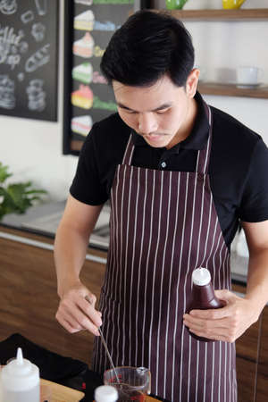 Smiling Asian barista young man pouring fresh Chocolate art in a plastic cup preparing for make Cappuccino iced coffee for according to order at counter bar in the morning at modern cafe.の写真素材