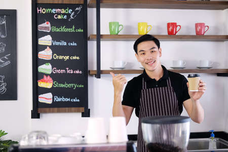 Smiling Asian barista young man holding a paper glass of hot coffee and Thumbs up for good taste and...