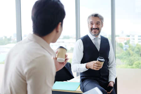 Smiling young Asian and Senior Caucasian businesspeople teamwork relaxing and drinking a mug of hot coffee together at modern office. Business and lifestyle concept.の写真素材