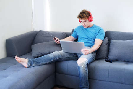 Young Caucasian man listening to musice by headphone and searching online social media with laptop....
