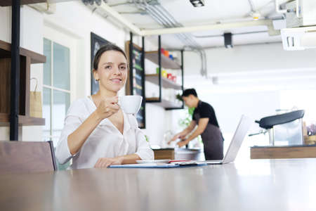 Smiling and successful Caucasian businesswoman is holding cup of hot coffee and online working and shopping in Social media with laptop in coffee shop. Business and technology concept.の写真素材