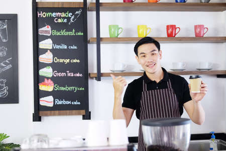 Smiling Asian barista young man holding a paper glass of hot coffee and Thumbs up for good taste and according to order at counter and menu bar in the morning at modern cafe.の写真素材
