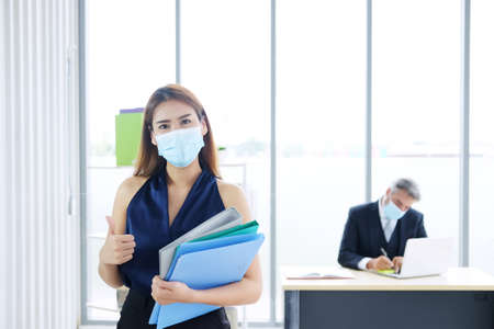 Smiling Young Asian businesswoman wearing mask preventing for Covid 19 virus holding Portfolio and document job. She Thumbs up for For good health of teamwork in manager room.の写真素材