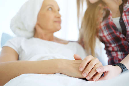 Smiling Caucasian daughter is Holding hands Elderly Mother in white headscarf is laying on bed in hospital after chemotherapy because she is suffering from cancer or Leukemia patient. focus on hands.の写真素材