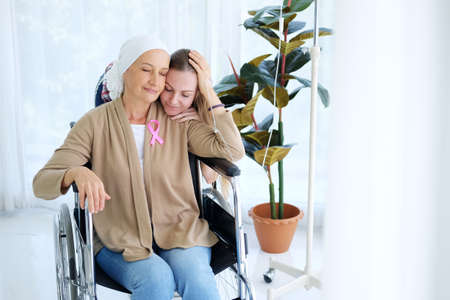 Caucasian young daughter Hug With love and care for her mother in white headscarf is sitting on wheelchair after chemotherapy. Elderly mother is suffering from cancer or Leukemia patient.の写真素材