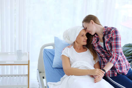Caucasian Elderly Mother in white headscarf is laying on bed in hospital after chemotherapy because she is suffering from cancer or Leukemia patient and young daughter is hugging and caring her mom.の写真素材