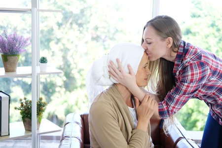 Smiling Caucasian young daughter is kissing on forehead à¹her Elderly Mother in white headscarf after chemotherapy because she is suffering from cancer or Leukemia patient.の写真素材