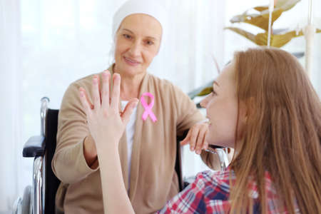 Smiling Caucasian young daughter Touch hands together with Elderly Mother in white headscarf sitting on wheelchair after chemotherapy. She is suffering from cancer or Leukemia patient. Focus on handsの写真素材