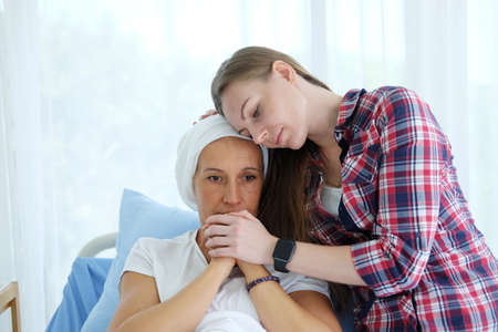Caucasian daughter is Holding hands Elderly Mother in white headscarf is laying on bed in hospital after chemotherapy because she is suffering from cancer or Leukemia patient. focus on hands.の写真素材