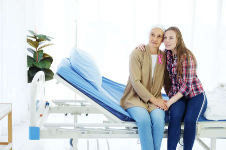 Smiling Caucasian Elderly Mother in white headscarf is sitting with young daughter on bed together in hospital after chemotherapy because she is suffering from cancer or Leukemia patient.の写真素材