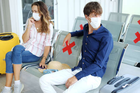 Serious Young Caucasian traveler couple love is wearing mask and sitting passenger waiting chairs for time of departure together because Plane delay in the airport. Honeymoon trip on vacation concept.の写真素材