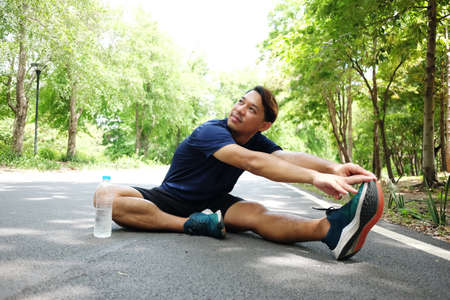 Smiling Asian man sitting on concrete floor for exercising and stretching the legs to relax the muscles after running and jogging in the garden. Sport and healthy Conceptの写真素材