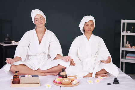 Caucasian and Asian woman in white bath towel for yoga meditation for create concentration and calm the mind on bed preparing for massage therapy at alternative medicine healing spa Center in Thailandの写真素材
