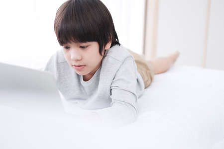 Smiling Asian little boy is studying and doing homework or playing game with laptop in bedroom. Homeschool and quarantine during Covid-19 virus epidemic concept. Distance Learning Online Educationの写真素材