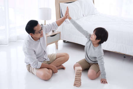 Smiling Asian father and cute little boy is enjoying and funny with wooden blocks game together in bedroom on holiday. Family activities and child educational for homeschool concept.の写真素材