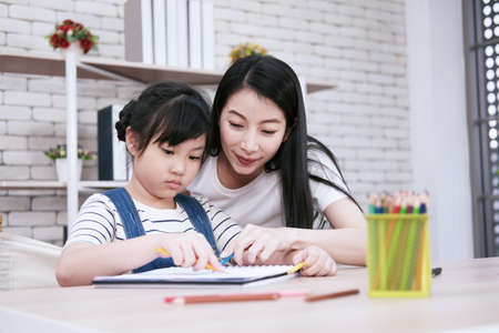 Smiling Asian mother and little asian girl child is drawing and Painting with wooden colored pencils on paper together in worksapce area at home. Homeschool and educational conceptの写真素材