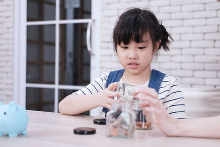 Smiling Asian little asian girl child is putting coins into piggy bank for saving money on wooden table. Child educational for homeschool concept.の写真素材