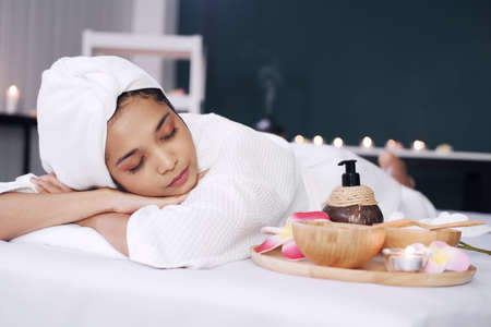 Smiling Asian woman in white bathrobe and headscarf and lie down and relaxing on bed preparing for massage therapy at alternative medicine healing spa Center in Thailandの写真素材