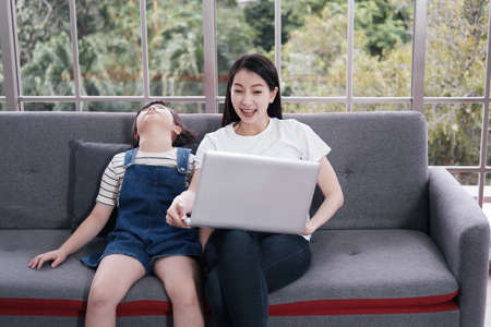 Smiling asian mother is searching and teaching little girl child for homework with laptop but her...