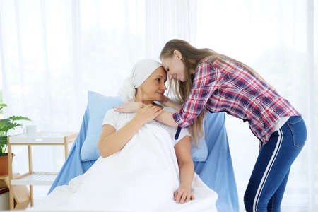 Caucasian daughter is kissing her Elderly Mother in white headscarf is sitting and smiling on bed...