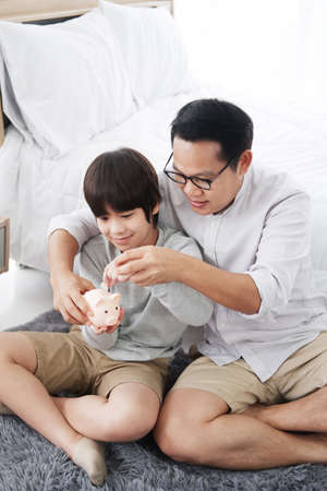 Smiling Asian little son child is putting coins into piggy bank for saving money for the future with father in bedroom. Child educational for homeschool concept.の写真素材