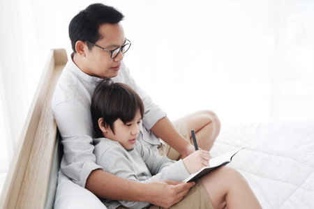 Smiling asian father and son child is writing a book and doing homework together on bed in bedroom