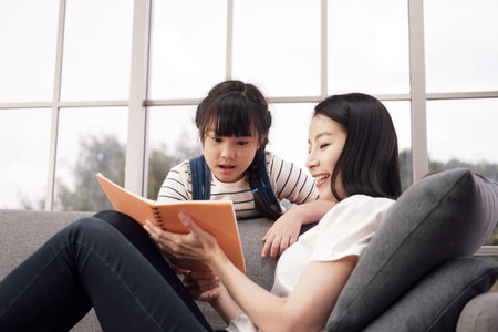 Smiling asian mother and little girl child is reading a book together on sofa in living room.の写真素材