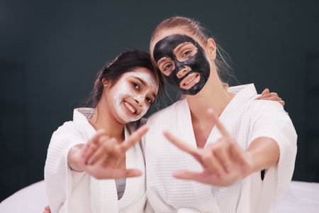 Caucasian and Asian woman in white bathrobe and black facial mask on face for treatment skin care together at spa.の写真素材