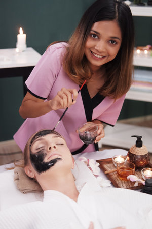 Professional Asian Thai Massage therapist woman is applying black facial mask for young caucasian...