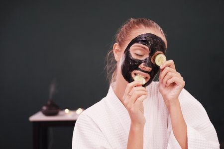 Smiling Caucasian woman in white bathrobe is holding cucumber slices and black facial mask on face for treatment skine care at spa.の写真素材