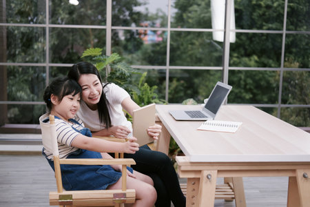 Smiling Asian mother and little girl child is selfie by tablet and online learning with laptop...