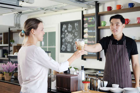 Smiling Asian barista young man serving a glass of iced latte coffee for Caucasian woman customer at counter bar in coffee shop.の写真素材