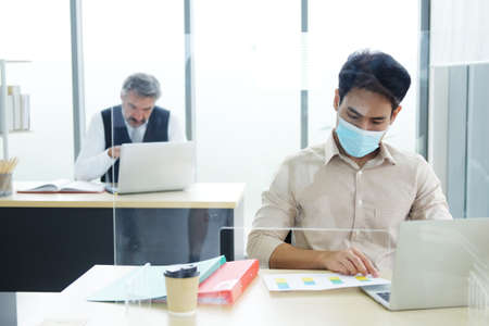 Caucasian boss and Asian businessman is wearing mask for preventing covid19 virus and partition with clear plastic on computer desk at office. Healthcare and New normal lifestyle in business concept.の写真素材