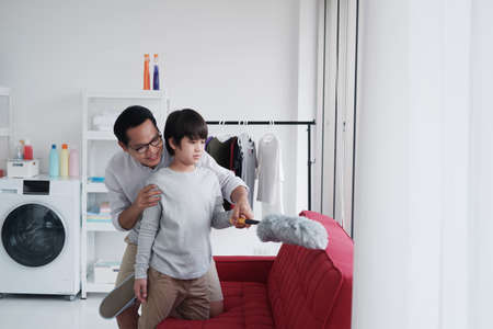 Smiling Asian father and little boy child is enjoying for House cleaning together in Laundry room. They is Dust off the red sofa  by Feather duster. Homeschool and Family time on holiday conceptの写真素材