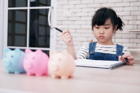 Smiling Asian little girl child is doing homework with piggy bank for saving money for the future on wooden table. Child educational for homeschool concept.の写真素材