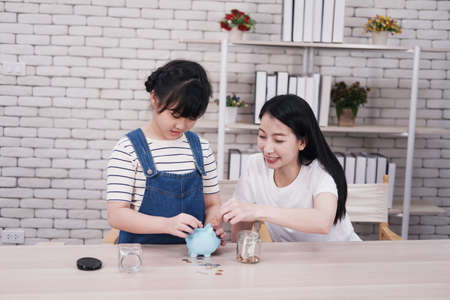 Smiling Asian little girl child is putting coins into piggy bank for saving money for the future with mother on wooden table. Child educational for homeschool concept.の写真素材