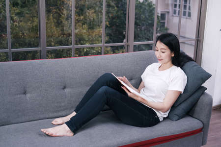 Smiling Asian businesswoman in white shirt is sitting on sofa and reading book in living room at home on holiday.の写真素材