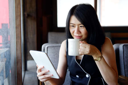 Smiling Asian businesswoman is drinking a cup of coffee and holding tablet for online shopping in...