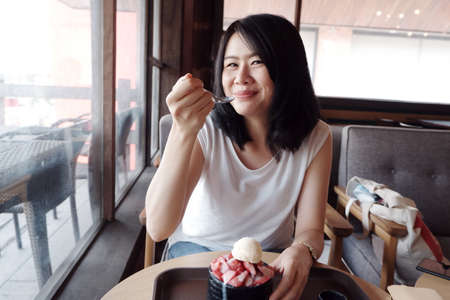 Smiling young Asian woman happy with delicious Strawberry Bingsu at coffee shop. Lifestyle and relaxtime of businesswoman on holiday conceptの写真素材