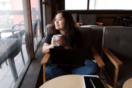 Smiling young Asian woman is drinking a cup of coffee and relaxing after online working with black...