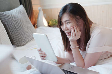 Smiling Asian young woman is laying on white bed holding tablet for online working and shopping with...