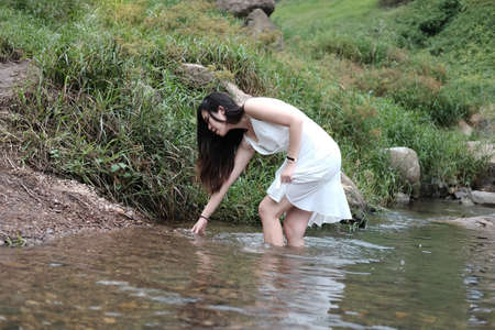 Smiling Beautiful asian female in white dress is standing on the rock and stream in the tropical...