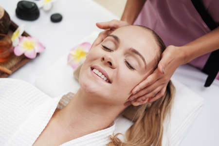 Asian Massage therapist woman is making traditional head and facial treatment massage to Caucasian female is relaxing and smilng on bed at alternative medicine healing spa Center in Thailand.の写真素材