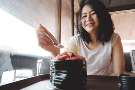 Smiling young Asian woman happy with delicious Strawberry Bingsu at coffee shop. Lifestyle and relaxtime of businesswoman on holiday concept.の写真素材