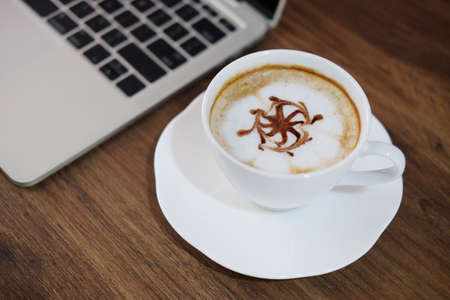 Latte art coffee cup with laptop computer on wooden table in cafe.