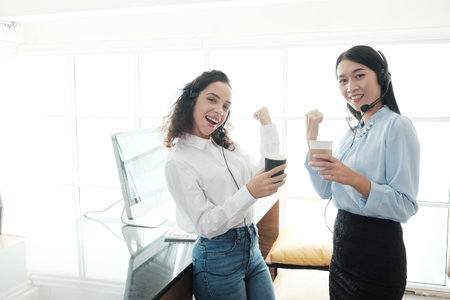 Smling asian and caucasian women is call center or secretary operator with headset and a microphone is relaxing and drinking coffee together after consultant to customers.の写真素材