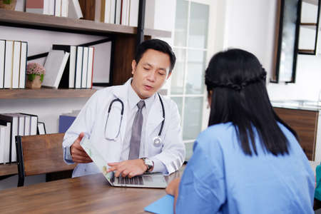 Asian chief physician man is meeting with surgeon doctor woman wears blue surgical gown. Medical team are talking and researching with laptop for treatment of patients in meeting room at hospital.の写真素材