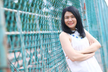 Beautiful asian woman in white dress smiling and standing with blue grating steel fence in natural park. Young Thai girl enjoy on holiday with sunlight in the garden.の写真素材