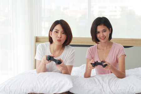Asian young sisters lovely couple on white bed hand holding joysticks and enjoy with game play...